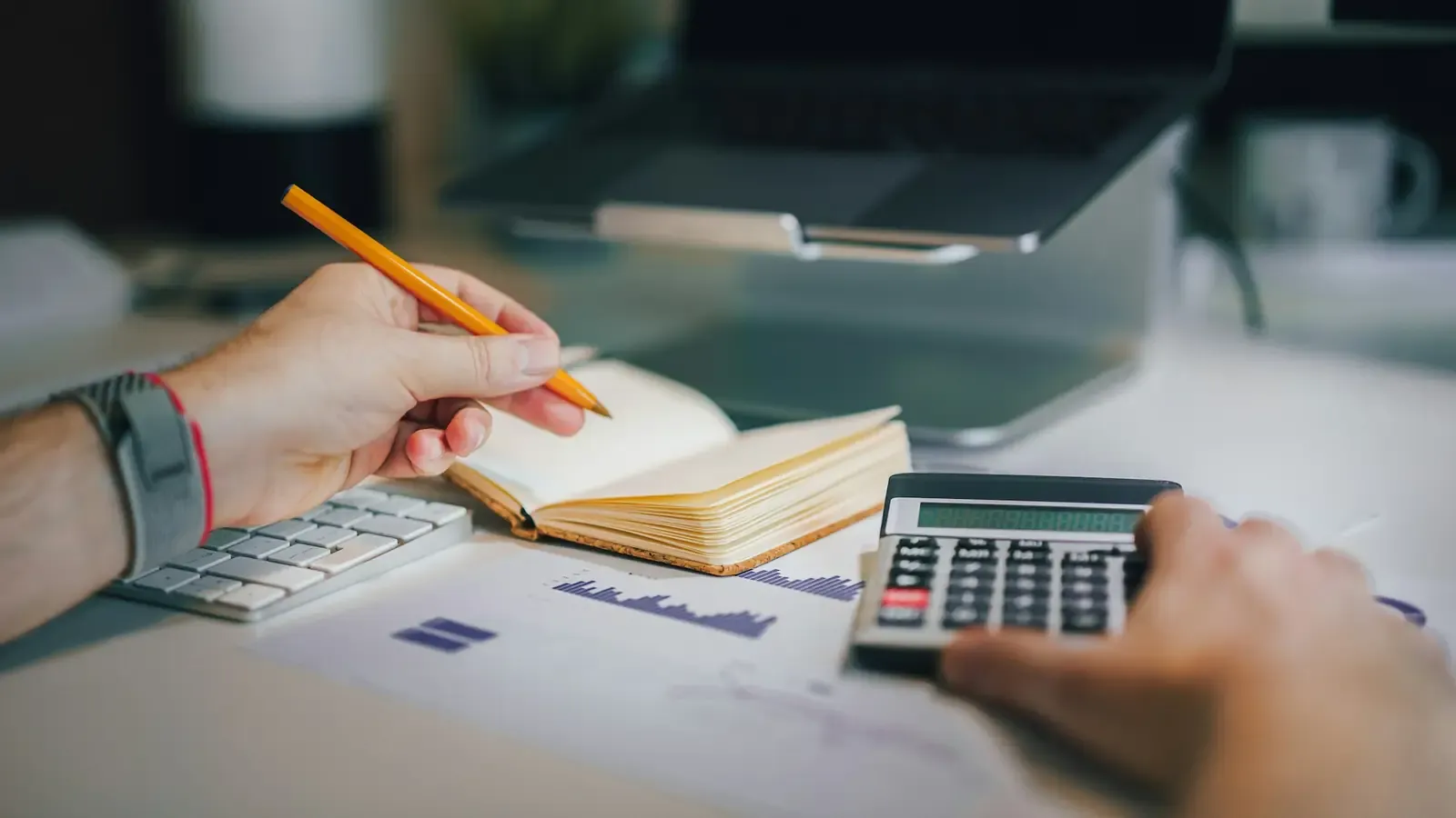Pessoa fazendo contas com uma calculadora e um caderno, com um notebook ao fundo sobre a mesa