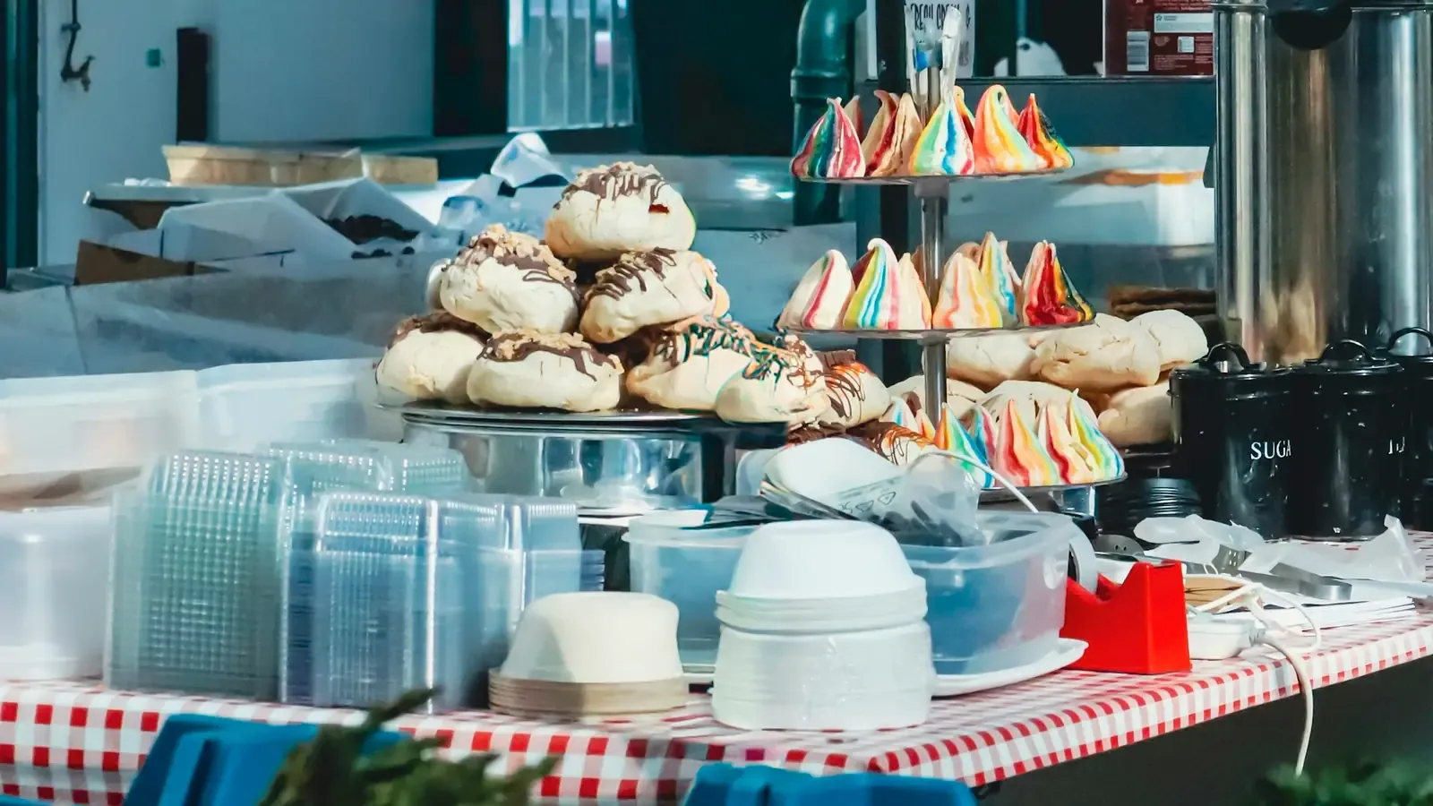 Banca de feira com merengues coloridos em suportes de dois andares, biscoitos com cobertura de chocolate e embalagens plásticas empilhadas sobre uma toalha xadrez vermelha