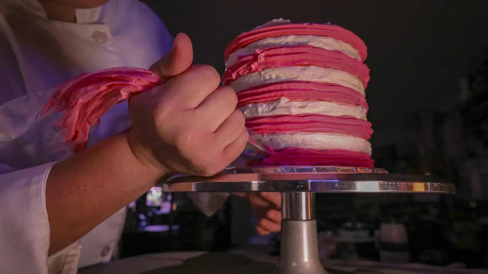 A baker piping the finish onto a pink striped layer cake on a turntable
