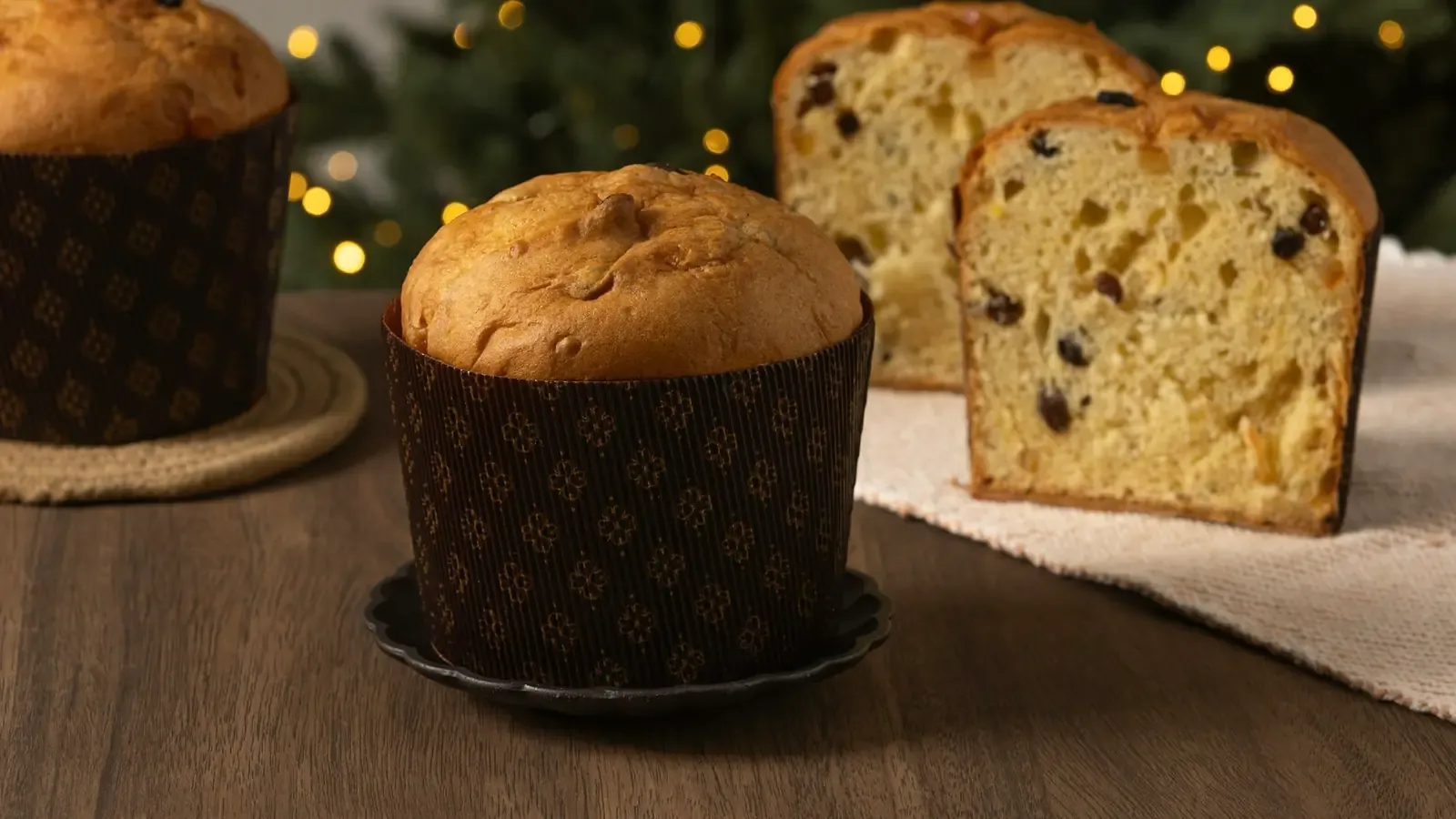 Panetone em forma de papel marrom na frente de outro panetone cortado, que mostra frutas cristalizadas e uvas-passas, com luzes de Natal desfocadas ao fundo