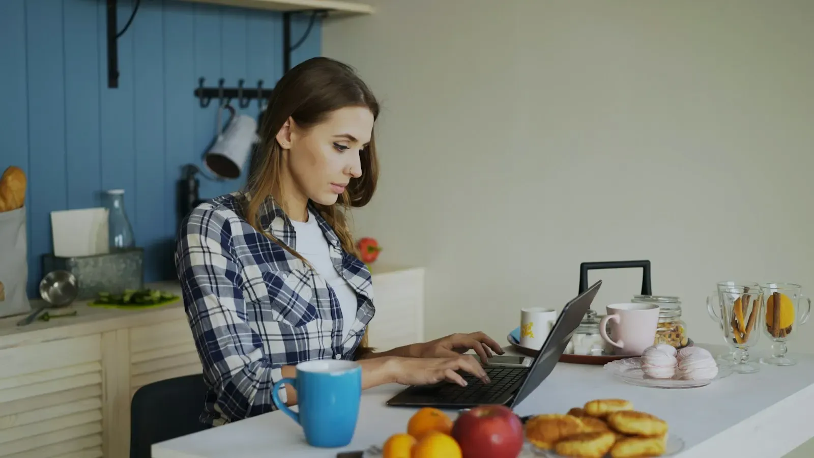 A woman working on a laptop at a kitchen table, with baked goods and eggs beside her